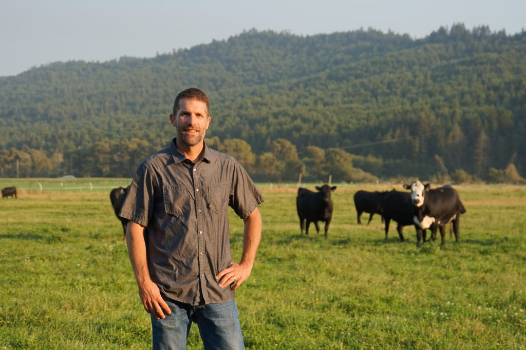 Mark Keller standing in a farm field with animals in the background.