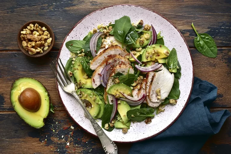 Spinach salad with grilled chicken fillet, avocado and walnuts on a plate over dark wooden background.