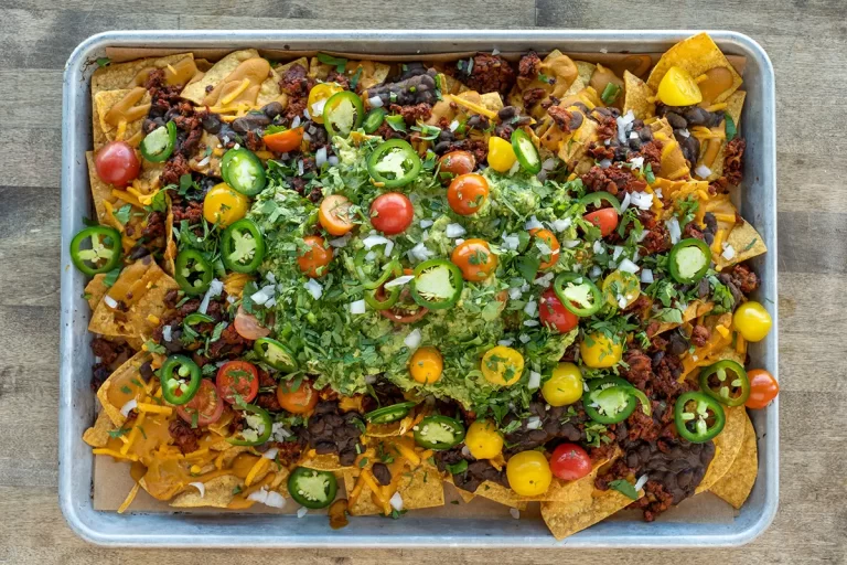 A sheet of vegan nachos loaded with guacamole, tomatoes, cashew cheese, cilantro, black beans and white onion.