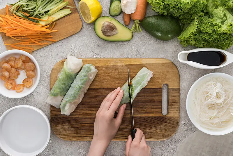 Top down view of someone cutting a spring roll on a cutting board with ingredients around it.