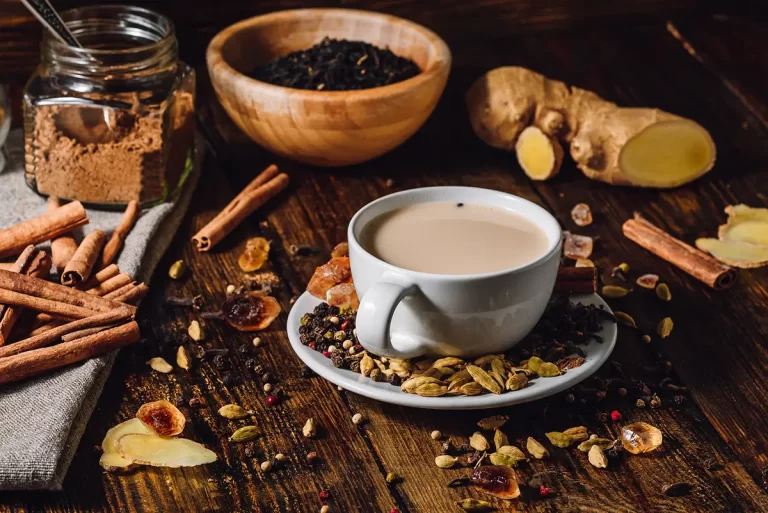 Masala Chai in a glass with raw ingredients, ginger, cinnamon and sugar on the table around it.