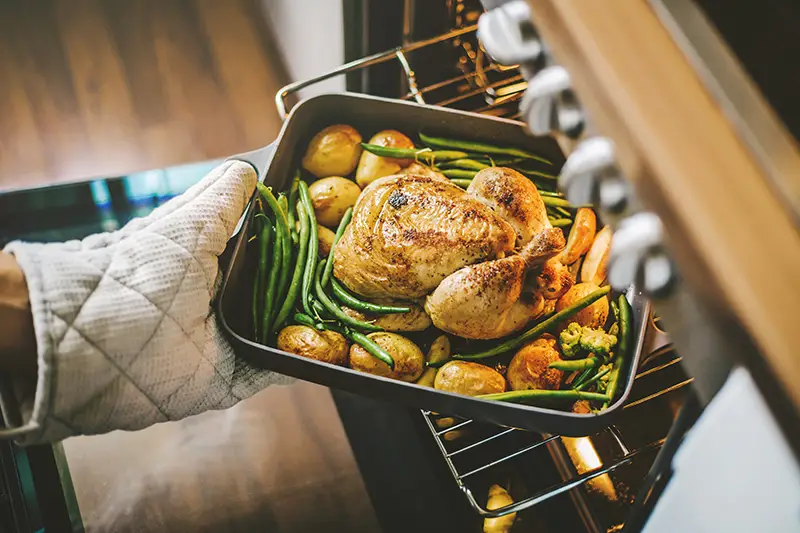 Fresh baked chicken and veggies in a roasting pan being pulled out of the oven by someone wearing oven mitts.