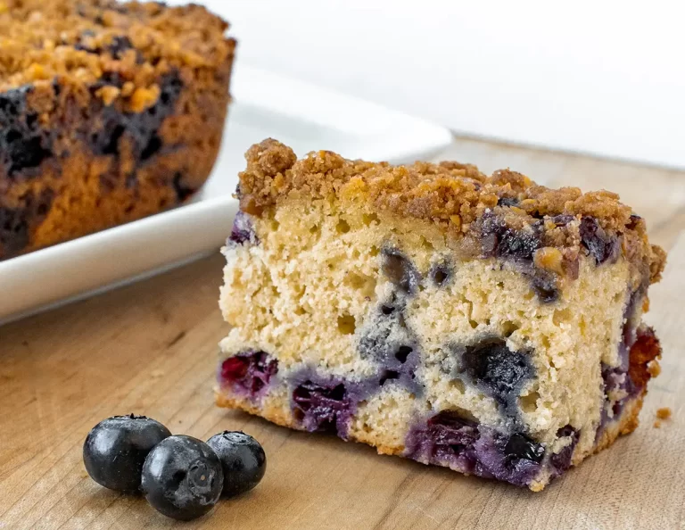 A slice of blueberry streusel cake on a cutting board with fresh blueberries in the foreground.