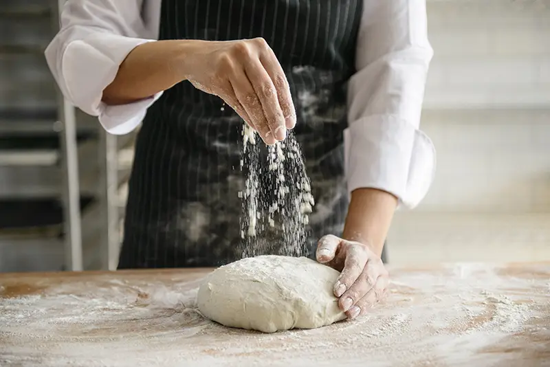 A person sprinkling flour on a ball of raw dough to make bread.